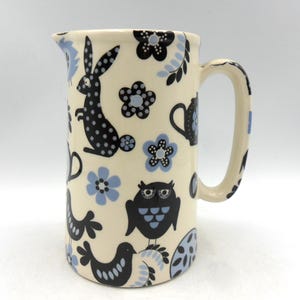 May include: A cream-coloured ceramic jug with a black and blue floral pattern. The pattern features owls, rabbits, and birds.
