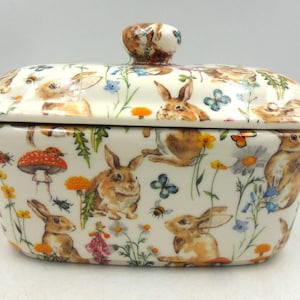 May include: A ceramic butter dish with a lid, decorated with a pattern of brown rabbits, flowers, butterflies, and insects on a white background. The lid has a small knob. The dish is rectangular with rounded edges.