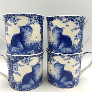 Könnte beinhalten: Vier gestapelte Keramikbecher, jeder mit einem blau-weißen Katzendesign. Die Becher haben einen floralen Rand und eine detaillierte Illustration einer Katze, die nach oben schaut. Die Becher sind weiß mit blauen Akzenten.