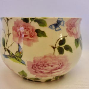 May include: A white ceramic bowl with a floral design featuring pink roses, green leaves, and a blue butterfly. The bowl has a rounded shape and a slightly flared rim.