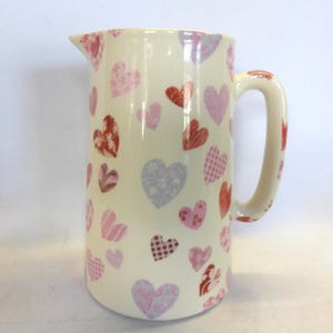 May include: A white ceramic jug with a handle, featuring a pattern of pink, red and purple hearts.