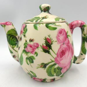 Peut inclure: Une théière en céramique de couleur crème ornée d'un motif répétitif de roses roses, de feuilles vertes et de boutons de rose. La théière comprend un couvercle assorti avec un petit bouton, un bec verseur incurvé et une anse robuste décorés.