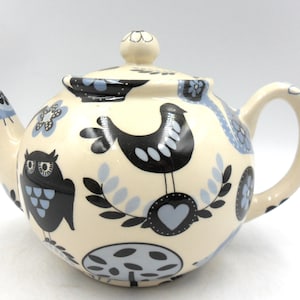 May include: Cream ceramic teapot with lid, spout, and handle. Decorated with black and light blue folk art designs, including an owl, a bird, flowers, and hearts. Designed for brewing tea, the teapot has a capacity of approximately 1.2 liters.