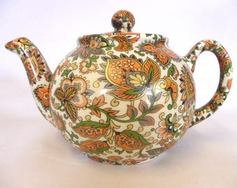Può includere: Una teiera in ceramica bianca con un motivo floreale in tonalità arancio, verde e grigio. La teiera ha un coperchio con un pomo in cima.
