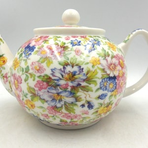 May include: A white teapot with a floral pattern in shades of pink, blue, and yellow. The teapot has a round body and a lid with a knob on top.