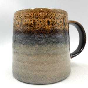 May include: A ceramic mug featuring a multi-toned glaze with a speckled tan and brown rim that transitions into a dark charcoal grey and fades into a light beige base. The mug has a smooth cylindrical body with a curved handle on the right side.
