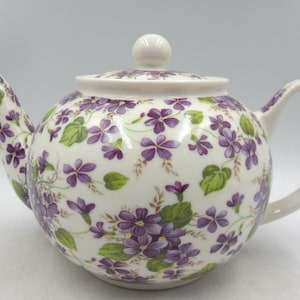 May include: A white teapot with a floral pattern of purple violets and green leaves. The teapot has a lid and a handle.