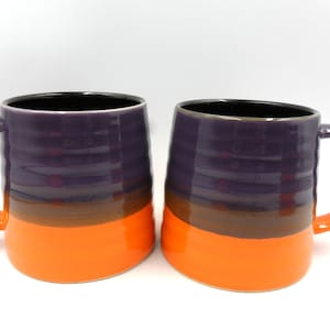 May include: Two ceramic mugs with a purple, brown, and orange colour scheme. The mugs have a rounded shape and a handle.