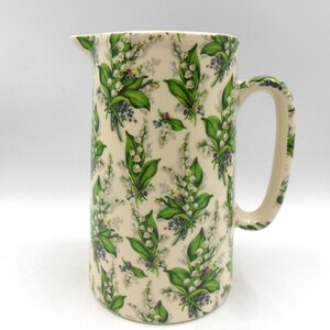 May include: A white ceramic jug with a floral pattern of green lily of the valley flowers and leaves. The jug has a wide mouth and a handle.