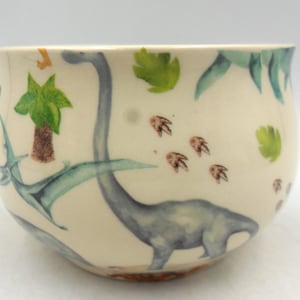 May include: A white ceramic bowl featuring a dinosaur and palm tree design. The bowl is decorated in shades of light blue, green, and brown.