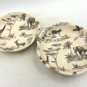 Könnte beinhalten: Zwei weiße Keramikschalen mit schwarz-weißen Safari-Tiermotiven. Die Schalen haben einen gewellten Rand und zeigen Giraffen, Elefanten und Zebras.