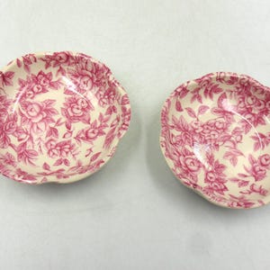 以下が含まれることがあります： ピンクの花柄の小さなクリーム色のボウルが2つ。ボウルはスカラップエッジで、ピンクのバラと葉の繰り返し模様で満たされています。ボウルはおそらくセラミックまたは磁器製です。