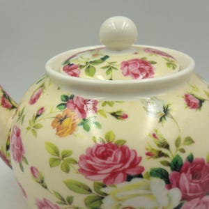 May include: A white ceramic teapot with a floral pattern featuring pink and yellow roses. The teapot has a lid with a knob on top.