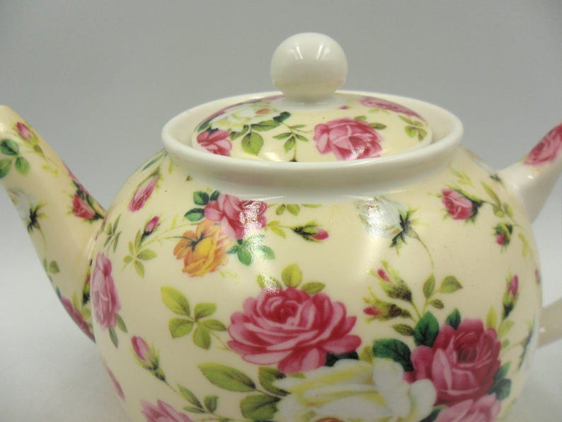 May include: A white ceramic teapot with a floral pattern featuring pink and yellow roses. The teapot has a lid with a knob on top.