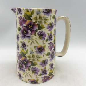 May include: A tall ceramic jug with a cream base, decorated with a floral pattern of purple pansies, green leaves, and white flowers. The jug features a curved handle and a pouring spout.