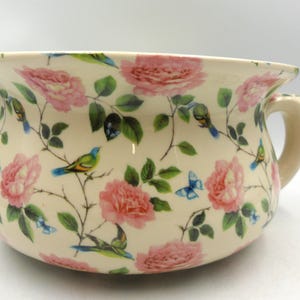 May include: A white ceramic chamber pot with a floral pattern featuring pink roses, green leaves, and blue birds. The pot has a handle on the side.