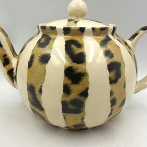 May include: A ceramic teapot featuring a repeating pattern of vertical cream-coloured stripes alternating with brown and black leopard print sections. The teapot includes a matching lid with a round knob, a curved spout, and a handle on the side.
