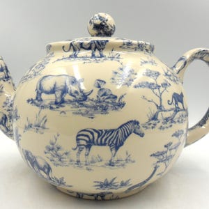 May include: A white ceramic teapot with a blue and white pattern of African animals, including zebras, rhinos, and elephants. The teapot has a round body and a spout on the left side. The handle is on the right side.