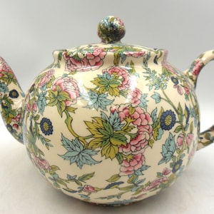 Peut inclure: Théière en céramique crème avec un motif floral de fleurs et de feuilles roses, bleues et vertes. La théière a un corps arrondi, un bec verseur incurvé et une anse en boucle. Le couvercle est surmonté d'un petit bouton à motifs floraux.