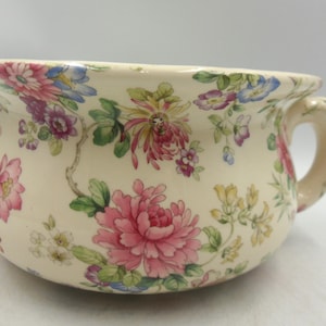 Könnte beinhalten: Cremefarbener Keramik-Nachttopf mit floralem Muster. Das Design zeigt rosa, lila, blaue und gelbe Blumen mit grünen Blättern. Er hat einen Griff und einen ausgestellten Rand.