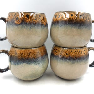 May include: Four ceramic coffee mugs stacked in pairs. The mugs have a rounded shape with a speckled brown and tan glaze at the top, transitioning to a blue and beige colour. Each mug has a black handle.