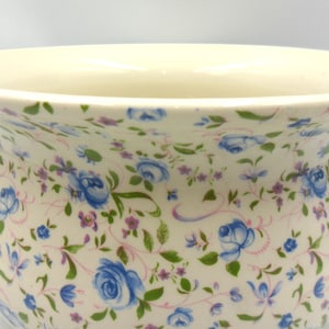 May include: A ceramic chamber pot with a cream-coloured base featuring a repeating pattern of blue roses, small purple flowers, and green leaves. The vessel has a wide, flared rim and a curved handle extending from the side of the rounded body.
