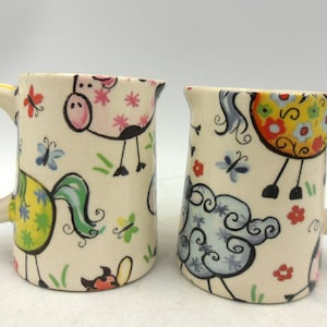 May include: Two cream ceramic mugs with quirky animal illustrations. One has a pink pig with a floral pattern, the other a blue sheep. Both mugs have a square handle and are decorated with butterflies and flowers, perfect for a farmhouse kitchen.