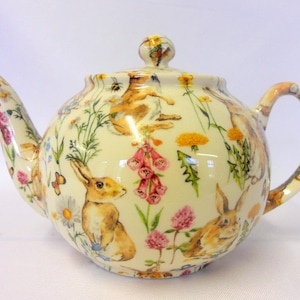 May include: A white ceramic teapot with a floral and bunny design. The teapot has a round body and a curved spout. The floral design features pink, yellow, and blue flowers. The bunnies are brown and white.
