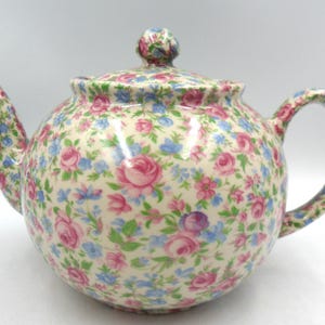 May include: A white teapot with a floral pattern in shades of pink, blue, and green. The teapot has a round body and a curved spout.