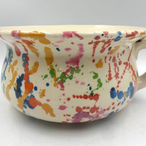 May include: A white ceramic chamber pot with a handle. The pot is decorated with colourful paint splatters in shades of orange, yellow, blue, green, pink and red.