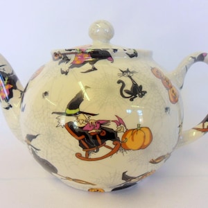 May include: A white ceramic teapot with a black handle and a black and white Halloween themed design. The teapot features witches, cats, pumpkins, and spiderwebs.