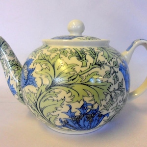 May include: A white teapot with a blue and green floral pattern. The teapot has a round body and a curved spout. The handle is attached to the side of the teapot.