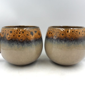 May include: Two ceramic mugs with a rounded shape and dark brown handles. The mugs feature a gradient design, transitioning from a light beige at the base to a dark brown speckled top. The mugs are designed for beverages.