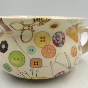 May include: A white ceramic mug with a handle, featuring a colourful pattern of buttons, needles, thread, and scissors. The mug is decorated with a sewing theme.