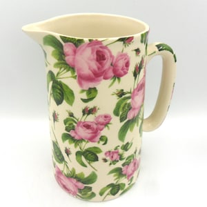 Könnte beinhalten: Eine weiße Keramikkanne mit einem Blumenmuster aus rosa Rosen und grünen Blättern. Die Kanne hat einen Henkel und eine breite Öffnung.
