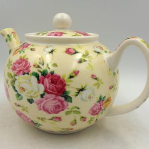 May include: A white ceramic teapot with a floral pattern of pink, white, and yellow roses. The teapot has a lid and a handle.