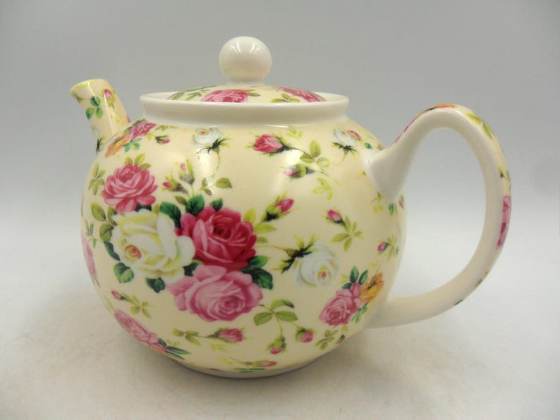 May include: A white ceramic teapot with a floral pattern of pink, white, and yellow roses. The teapot has a lid and a handle.