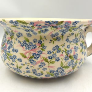 May include: A ceramic chamber pot featuring a cream-coloured base decorated with a repeating pattern of small blue flowers, green leaves, and thin pink ribbons. The vessel has a wide, flared rim and a single sturdy handle on the right side.