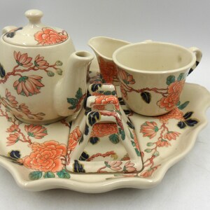 Könnte beinhalten: Ein cremefarbenes Vintage-Teeservice mit Teekanne, Sahnekännchen, Zuckerdose und Tasse, alles auf einem passenden Tablett. Das Set ist mit orangefarbenen und rosa Blumenmustern und grünen Blättern verziert.