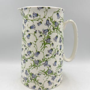 May include: A white ceramic jug with a floral pattern of blue and green bellflowers. The jug has a handle and a wide mouth.