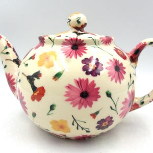 May include: A white ceramic teapot with a floral pattern. The teapot features a pink, purple, and yellow floral design with green stems and leaves. The teapot has a round body and a curved spout.