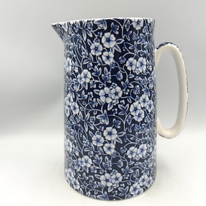 May include: A ceramic jug featuring a dark blue background with a repeating white floral pattern of small blossoms and leaves. The vessel has a curved handle on the right side and a pouring spout at the top rim, with a solid white interior.