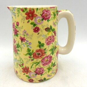 May include: A yellow ceramic jug with a white handle and a floral pattern featuring pink, red, and white flowers.
