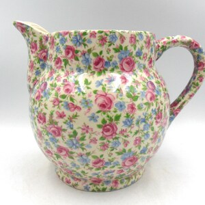 May include: A white ceramic jug with a floral pattern in shades of pink, blue, and green. The jug has a wide base and a rounded body. It features a handle on the side.