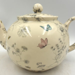 May include: Cream ceramic teapot with a lid, handle, and spout. The teapot is decorated with delicate grey floral and butterfly patterns in pink and blue. The teapot has a round shape.