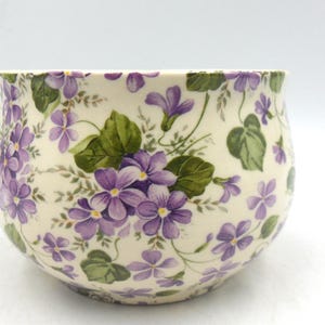Peut inclure: Un bol en céramique blanche avec un motif floral. Le bol est décoré de fleurs et de feuilles violettes et vertes.