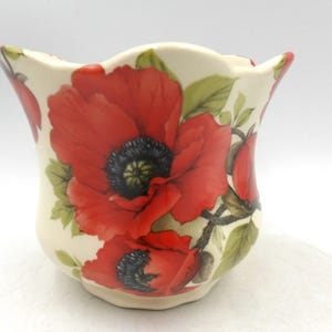 May include: Cream ceramic vase with a scalloped edge, featuring bold red poppy flowers and green foliage. The poppies have black centres, creating a striking contrast. A decorative floral accent for the home.
