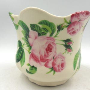 May include: A white ceramic planter with a scalloped rim features a floral design of pink roses and green leaves.