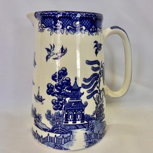 May include: A white ceramic jug with a blue Willow pattern design. The jug features a spout, a curved handle, and a detailed landscape scene with a pagoda, trees, and birds. The rim has a blue floral border.