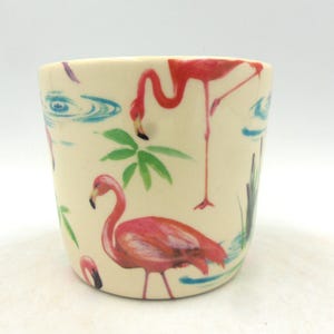 Peut inclure: Une tasse en céramique blanche avec un motif de flamants roses. La tasse a un design aquarelle bleu et vert d'eau et de feuilles de palmier.